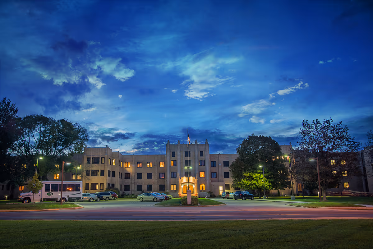 Exterior view of a large multi-story senior living facility building at dusk with lights on inside. The building is surrounded by trees and a parking lot with several cars and a shuttle bus parked. The sky is partly cloudy with a deep blue color.