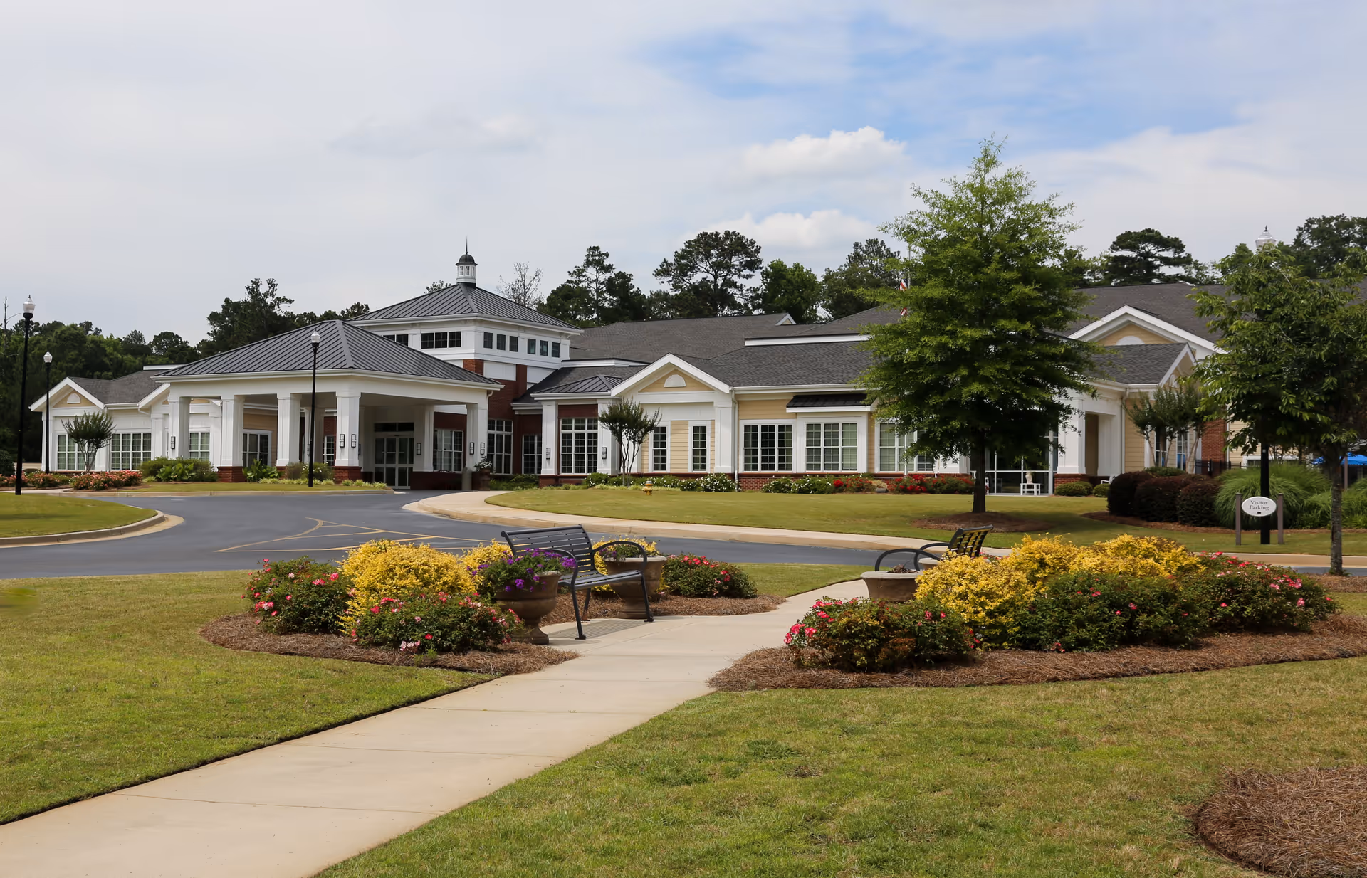 Front view of a single-story health and rehabilitation building with a porte-cochère, circular driveway, landscaped flower beds and benches.