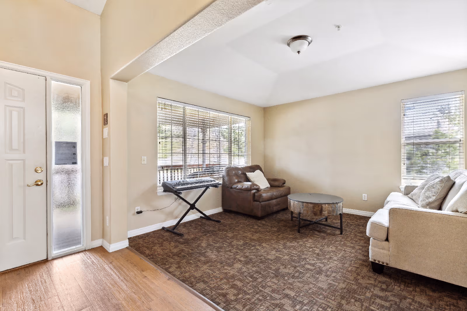 A cozy living room with beige walls and carpeted floor. The room features a brown leather armchair with a white pillow, a beige sofa with multiple cushions, and a round wooden coffee table. There is a keyboard on a stand placed near a large window with blinds, allowing natural light to brighten the space. The entrance door with frosted glass panels is visible on the left side.
