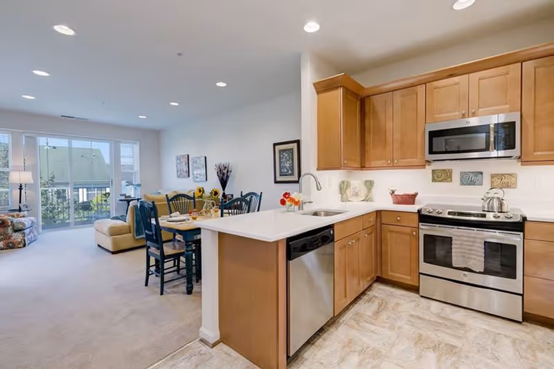 Bright open kitchen and living area in a senior living facility. The kitchen features wooden cabinets, a stainless steel dishwasher, oven, and microwave. A white countertop island with a sink separates the kitchen from the dining area, which has a table set with chairs. The living area has a sofa, armchair, and large windows letting in natural light.