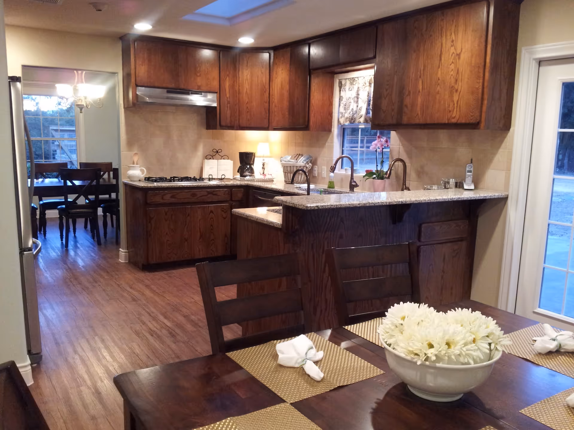 Interior view of a kitchen and dining area with wooden cabinets, granite countertops, a gas stove, a coffee maker, and a dining table set with placemats, napkins, and a bowl of white flowers. There is a window with a floral curtain above the sink and a glass door leading outside.