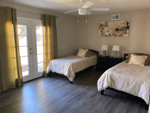 A bedroom with two single beds, each with beige bedding and pillows. Between the beds is a black nightstand with two matching lamps. Above the nightstand hangs a floral painting. The room has light-colored walls, wood flooring, a ceiling fan, and French doors with green curtains letting in natural light.
