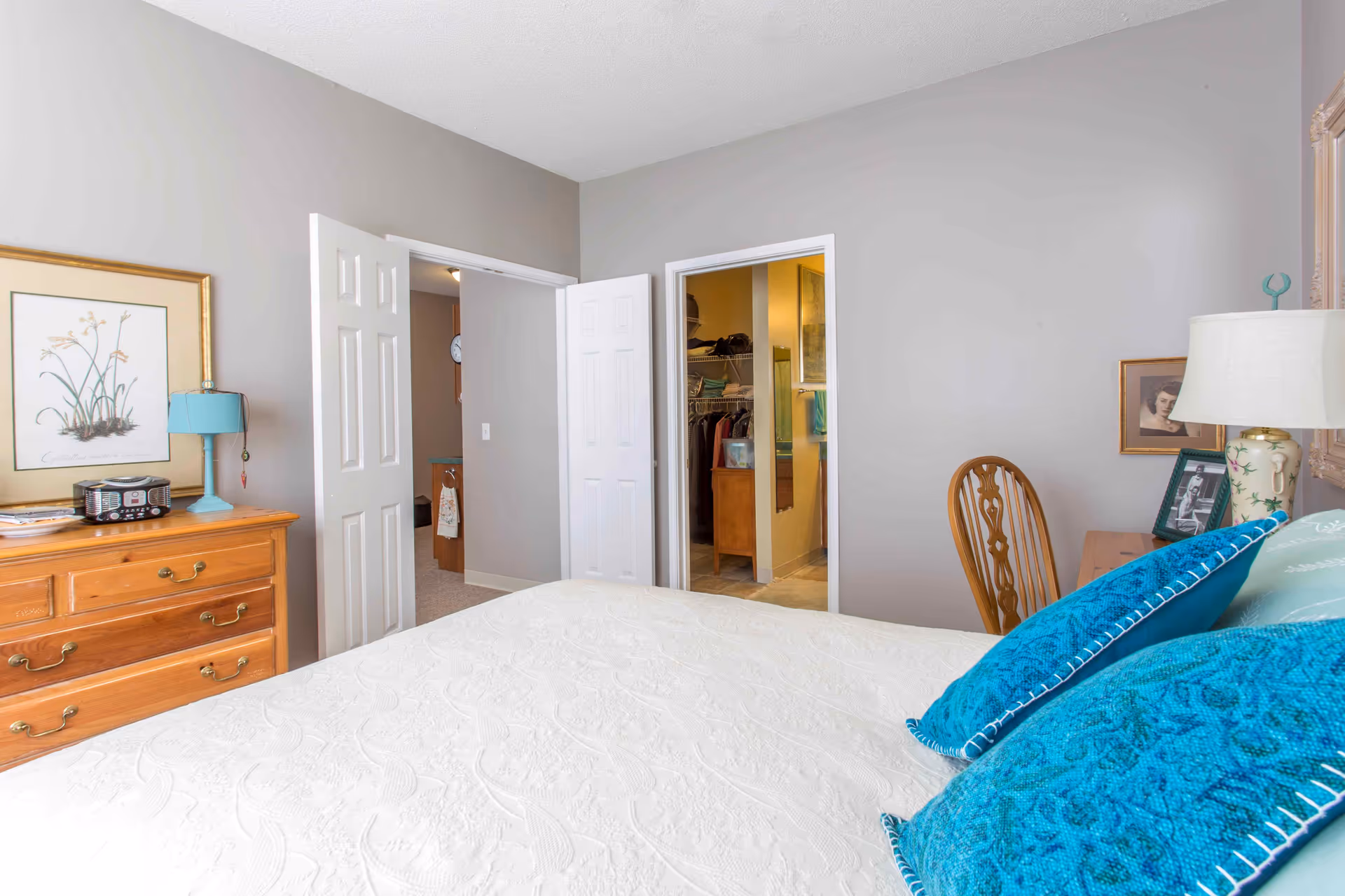 Bright bedroom interior showing a bed with a white coverlet and teal pillows, a wooden dresser with a lamp and artwork, and open doors revealing a closet and hallway.
