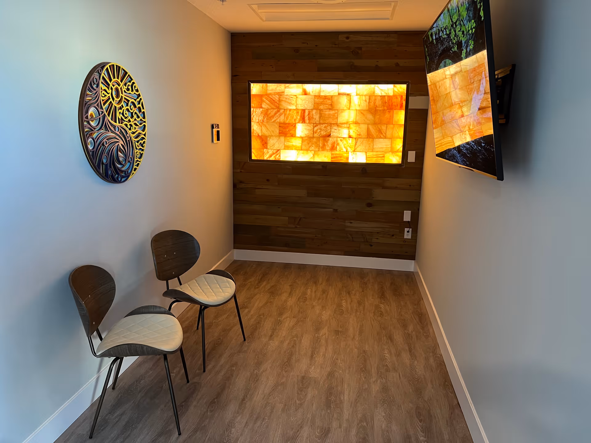 A small room with two modern wooden chairs with white cushions placed against a light blue wall. On the opposite wall, a flat-screen TV is mounted. The far wall features a wooden panel with a backlit decorative panel emitting a warm orange glow. The floor is covered with wood-patterned vinyl or laminate.