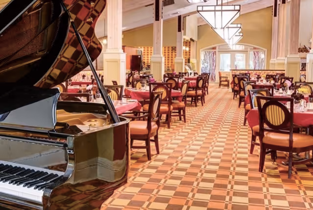Interior view of a dining room with multiple tables covered in red tablecloths and set with glasses and napkins. A grand piano is visible in the foreground on the left side. The room has a patterned carpet and large windows at the far end letting in natural light.