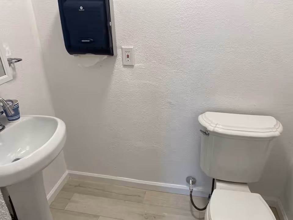 A small bathroom with a white pedestal sink on the left and a white toilet on the right. Above the sink is a blue paper towel dispenser mounted on the wall. The walls are white with a textured finish, and the floor has light-colored wood-like tiles.