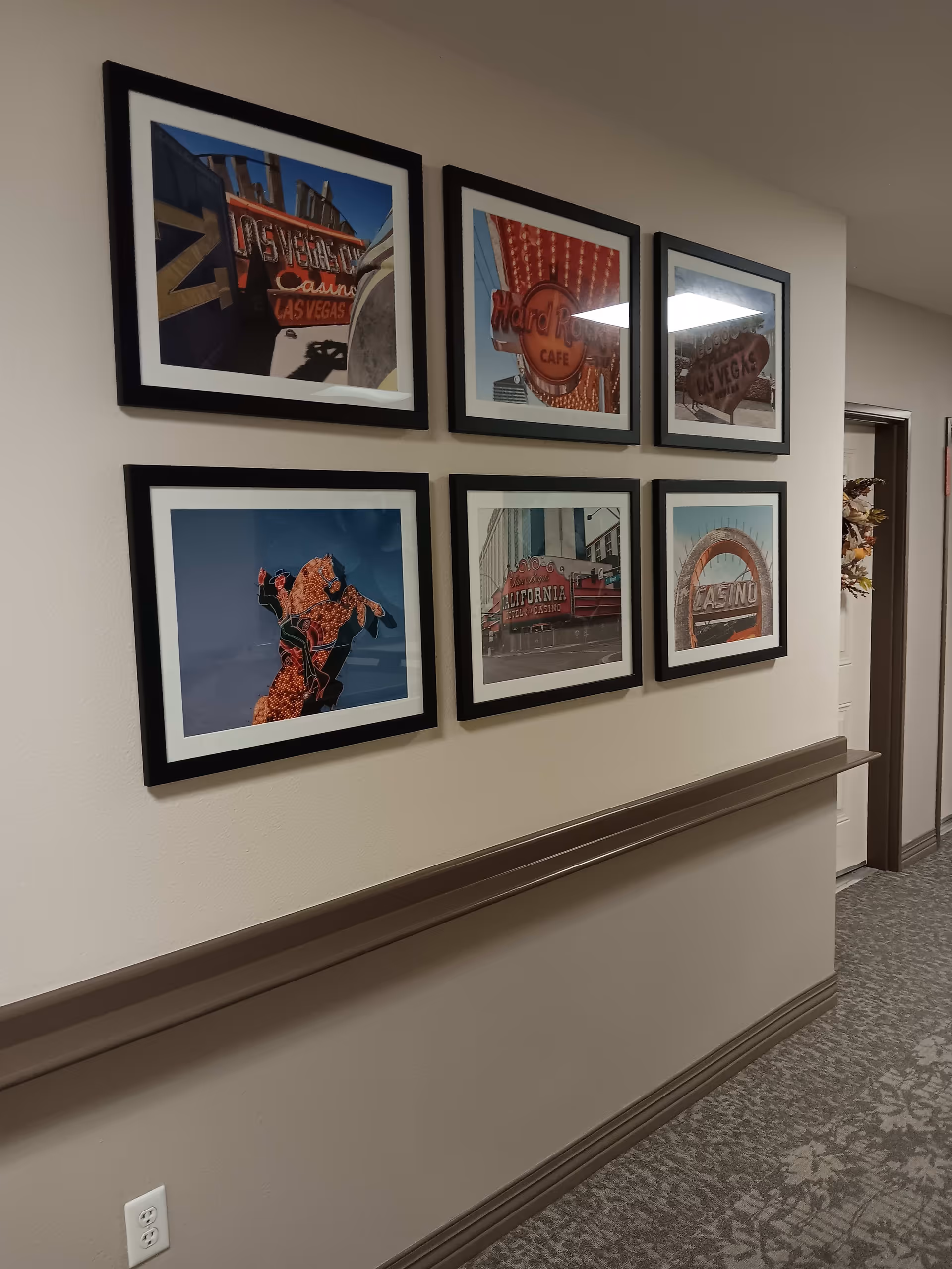 A hallway wall decorated with six framed photographs of vintage Las Vegas casino signs. The wall is painted beige with a brown chair rail and baseboard. The carpet has a floral pattern, and there is a doorway visible at the end of the hallway.
