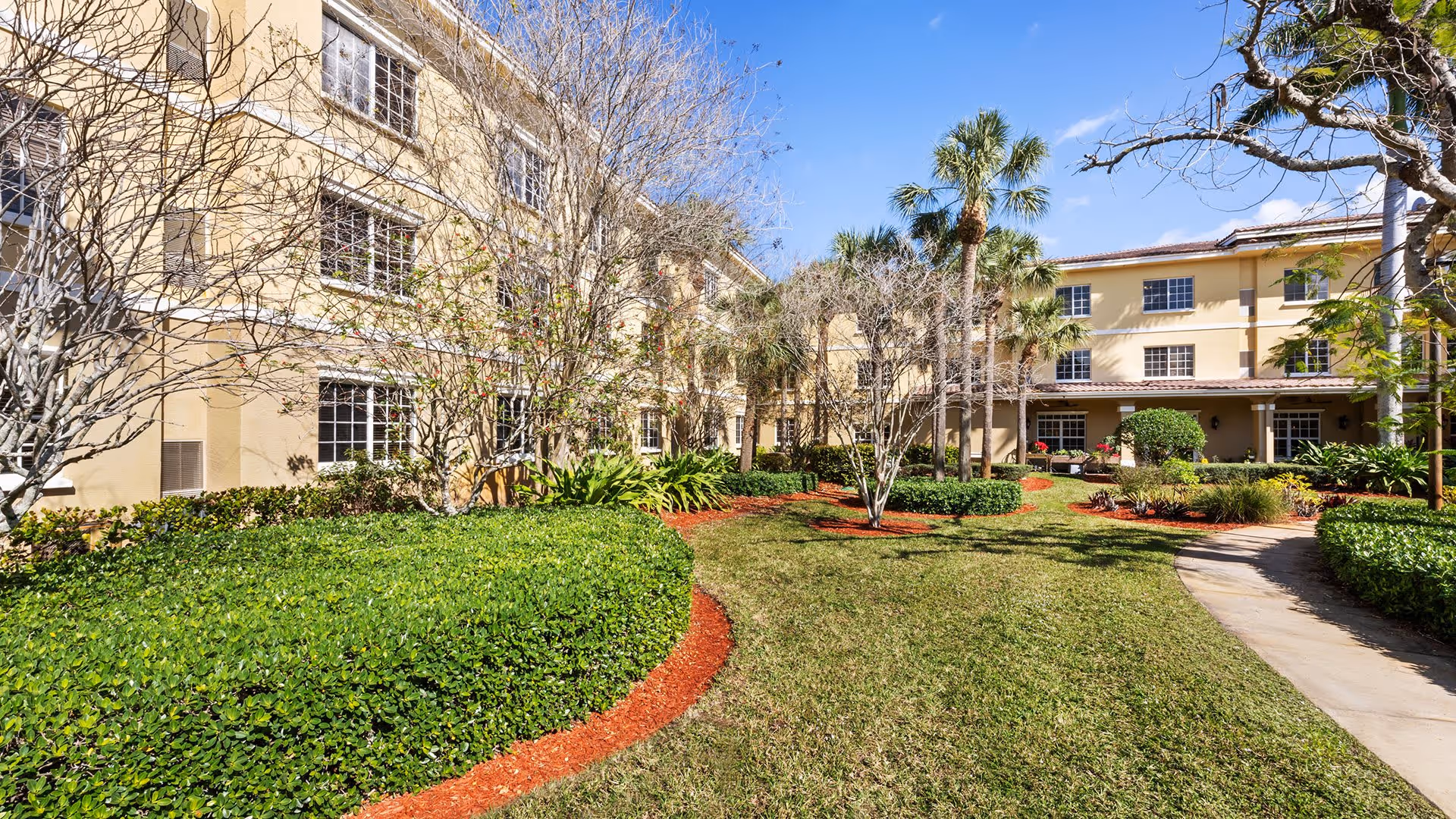 Outdoor courtyard area of a senior living facility with well-maintained green grass, various shrubs, palm trees, and other plants. The courtyard is surrounded by a beige multi-story building with multiple windows. A paved walkway curves through the garden area under a clear blue sky.