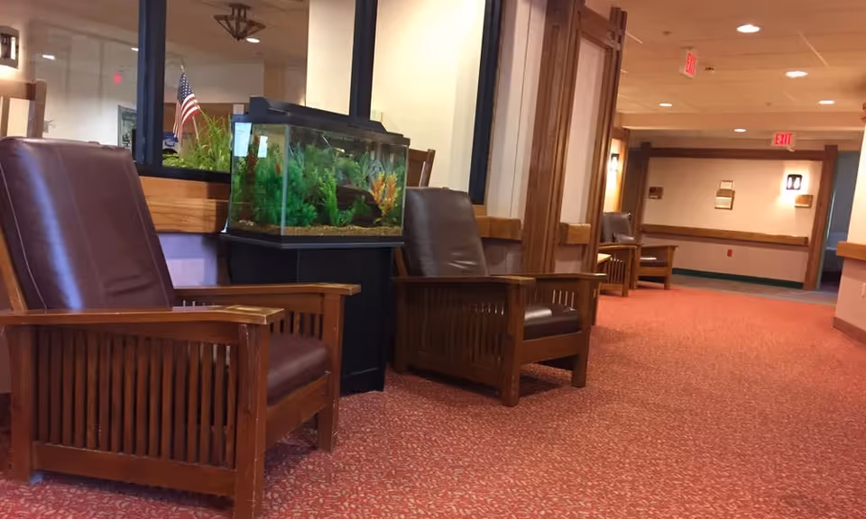 A hallway in a senior living facility with red patterned carpet, wooden armchairs with brown cushions, and a fish tank on a black stand. The walls are light-colored with wooden trim, and there are exit signs and wall-mounted lights visible.