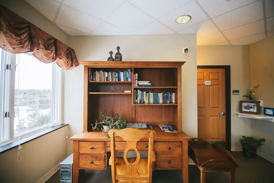 A wooden desk with a matching chair and hutch filled with books is positioned against a beige wall. On the desk are a keyboard, mouse, a plant, and some magazines. To the left, there is a large window with a decorative valance, letting in natural light. To the right, a wooden door and a small shelf with framed pictures and a plant are visible.