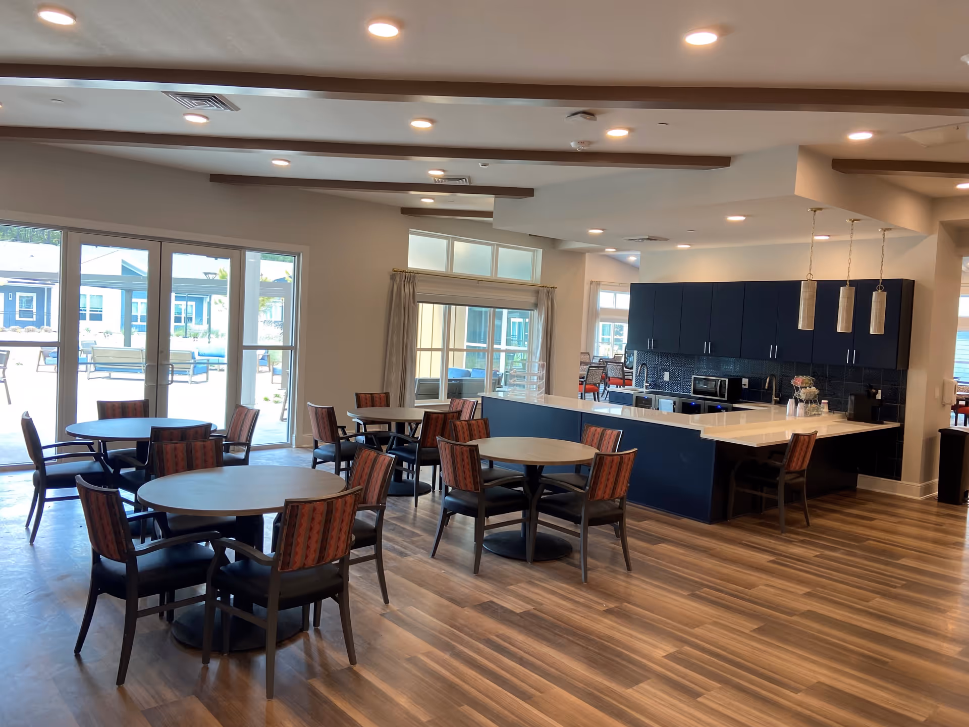 A bright and spacious dining area with several round tables and chairs arranged neatly on a wood-patterned floor. Large windows and glass doors let in natural light and offer a view of an outdoor seating area. To the right, there is a modern kitchen with dark blue cabinets, a white countertop island, and three pendant lights hanging above it.