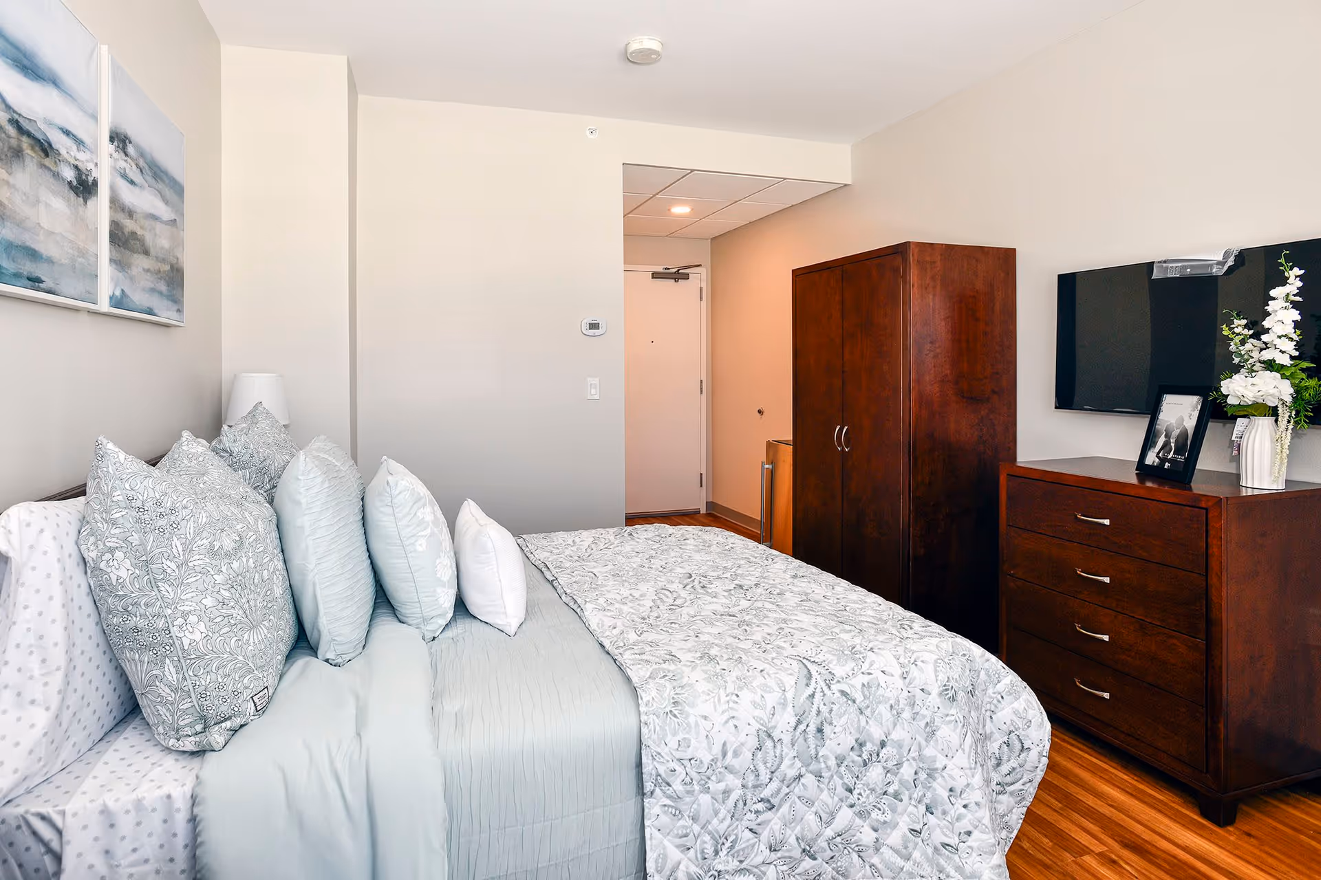 A neatly made bed with multiple pillows in light blue and white patterns in a bedroom. The room features light-colored walls, wooden flooring, a wooden wardrobe, a wooden dresser with a vase of white flowers and a framed photo, and a wall-mounted flat-screen TV. Two abstract paintings hang on the wall above the bed.