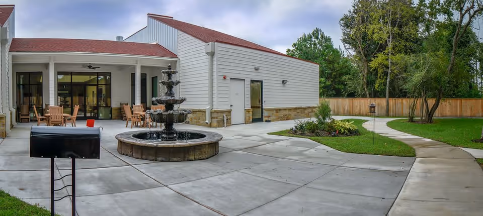 Outdoor courtyard area of Village Green Memory Care Community Cypress (Louetta) featuring a multi-tiered water fountain in the center, surrounded by concrete walkways and green lawn with trees and shrubs. There are wooden chairs and tables under a covered patio attached to a white building with a red roof.