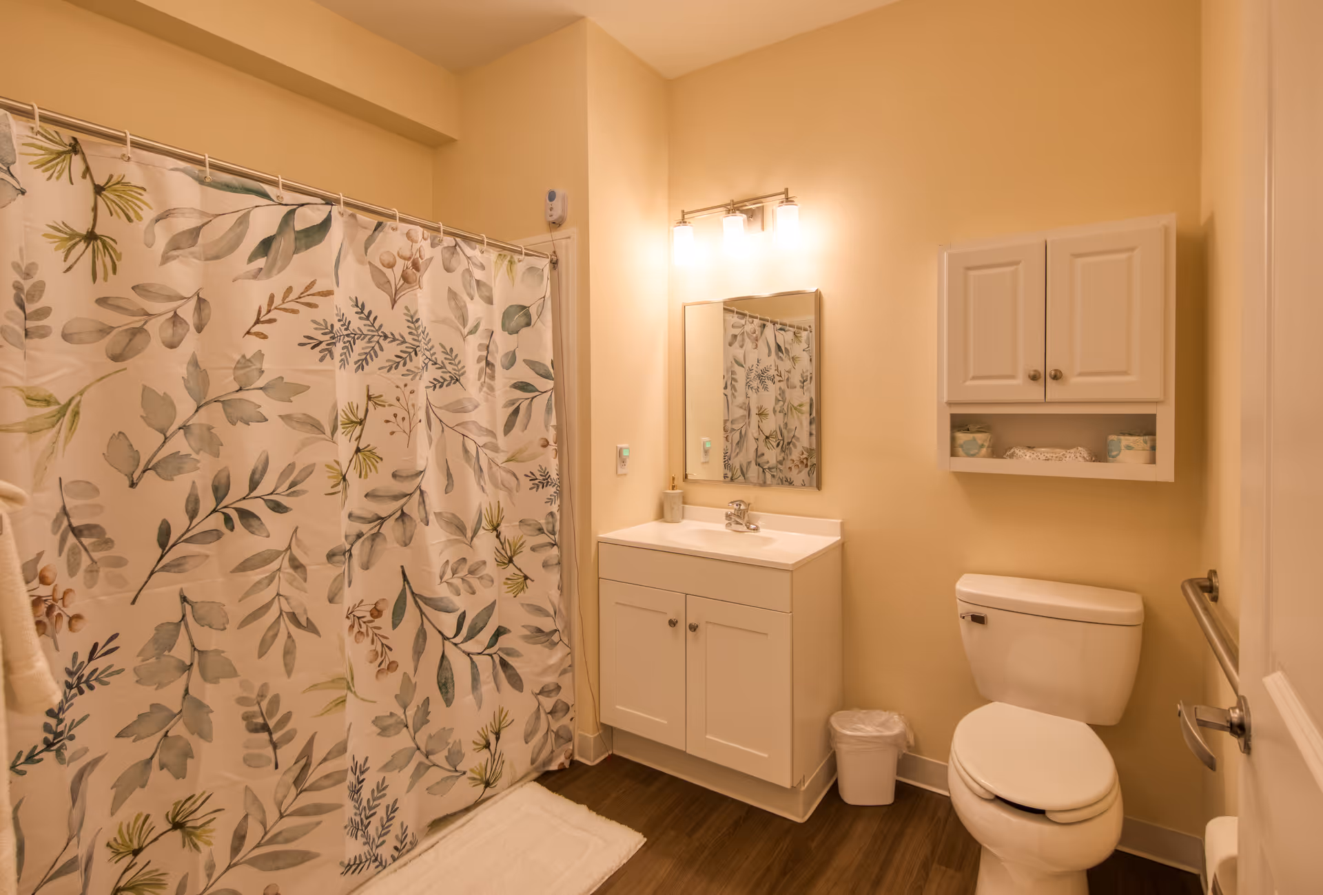 A clean and well-lit bathroom featuring a white toilet, a white vanity with a sink and mirror above it, a wall-mounted cabinet with toiletries, and a shower with a floral-patterned curtain. The floor is wood-style, and the walls are painted a soft beige color.