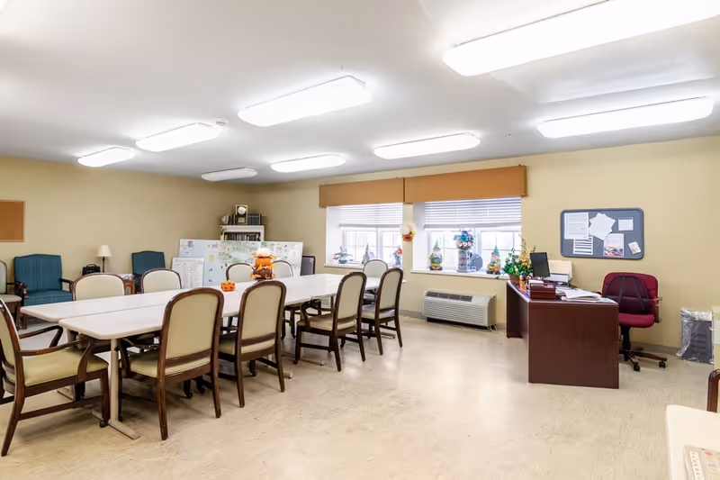 A well-lit room with a long rectangular table surrounded by chairs. The room has beige walls, two windows with blinds, and a desk with a computer and office chair in one corner. There are decorations on the windowsill and a bulletin board on the wall. Additional seating is arranged along the walls.