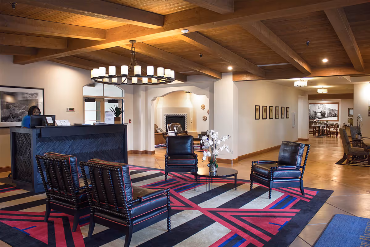 Spacious reception lobby with a front desk, leather chairs around a coffee table on a colorful geometric rug, and exposed wooden ceiling beams.