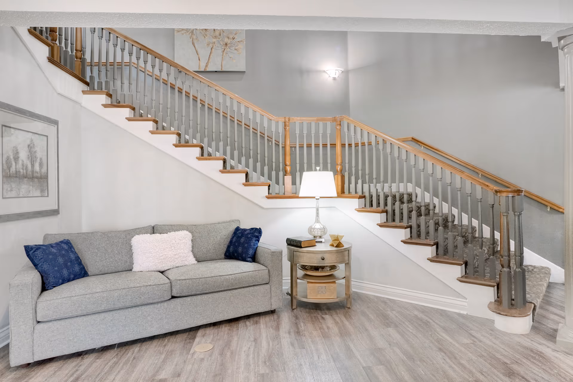 A cozy living area featuring a gray sofa with two blue pillows and one white fluffy pillow, a round wooden side table with a glass lamp, books, and decorative items, positioned under a staircase with wooden handrails and gray balusters. The walls are painted light gray and decorated with framed artwork.