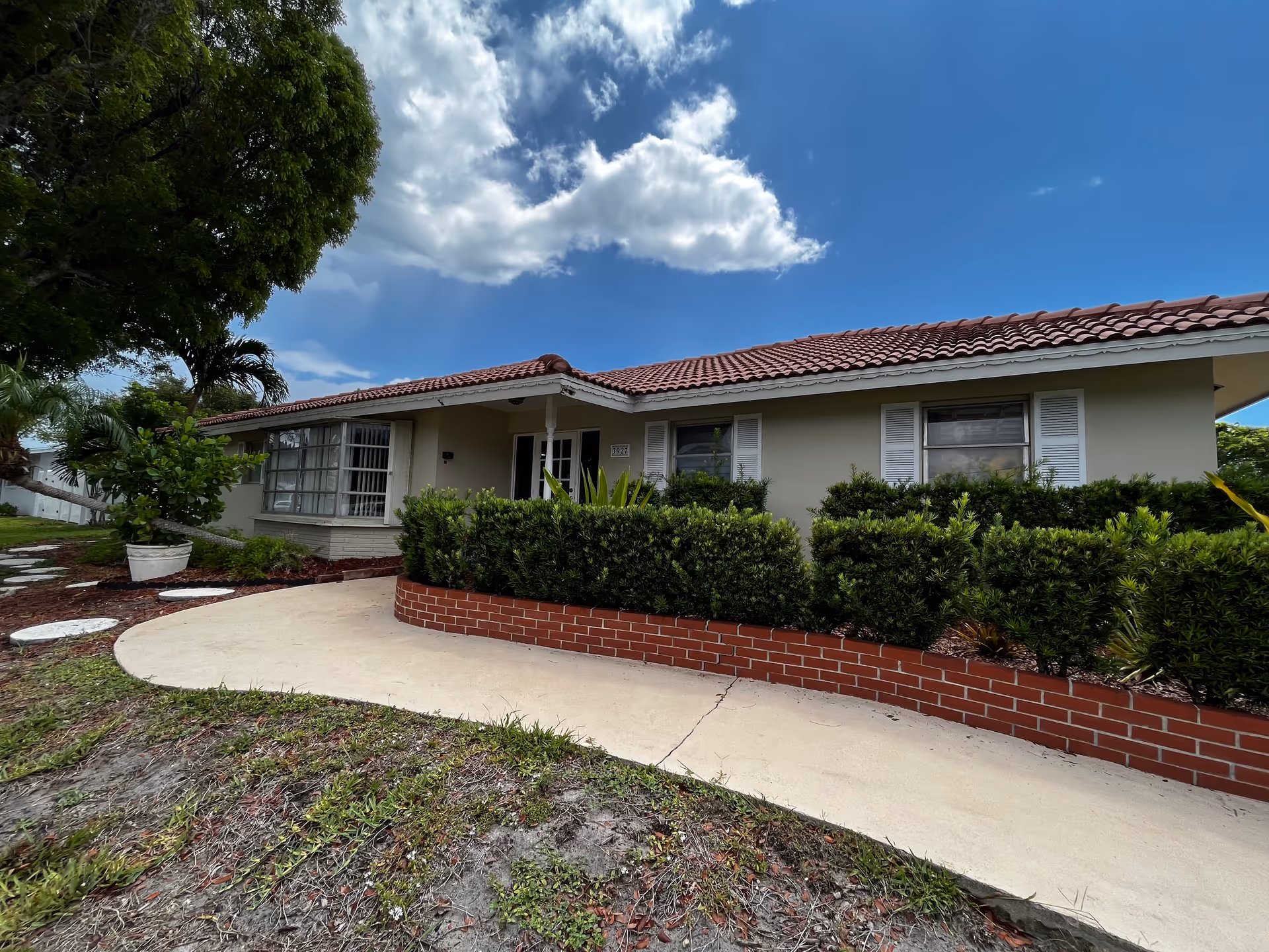 Single-story house with a red tile roof, trimmed shrubs and a curved concrete walkway under a partly cloudy sky.