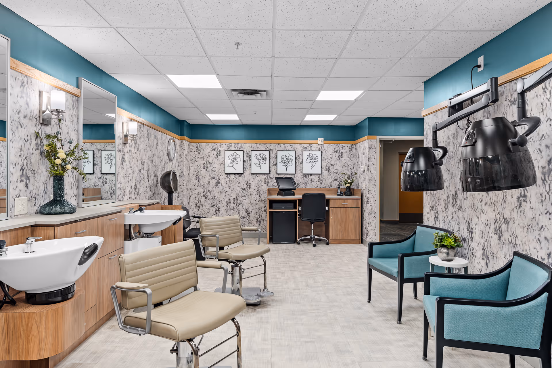 A modern hair salon area inside a senior living facility with two beige salon chairs in front of sinks, two black hair dryers mounted on the wall, and two teal armchairs with a small white table between them. The walls have a textured gray and white pattern with teal accents near the ceiling. A wooden desk with a computer and chair is visible in the background, along with framed floral artwork on the wall.