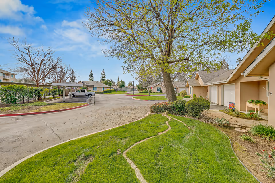 Outdoor view of a senior living facility showing a curved driveway with parked vehicles, green grass, trees, and beige-colored buildings with garages under a partly cloudy blue sky.