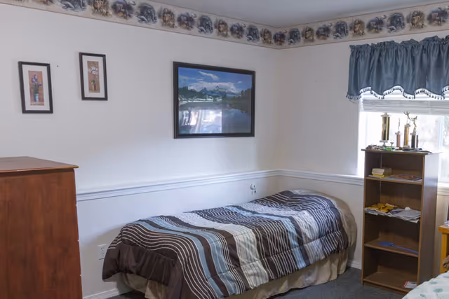 A small bedroom with a single bed covered in a striped brown, blue, and beige comforter. The walls are white with a decorative border near the ceiling featuring animal designs. There are three framed pictures on the wall above the bed, and a wooden dresser is partially visible on the left. A wooden bookshelf with trophies and books is next to a window with blue curtains on the right side of the room.
