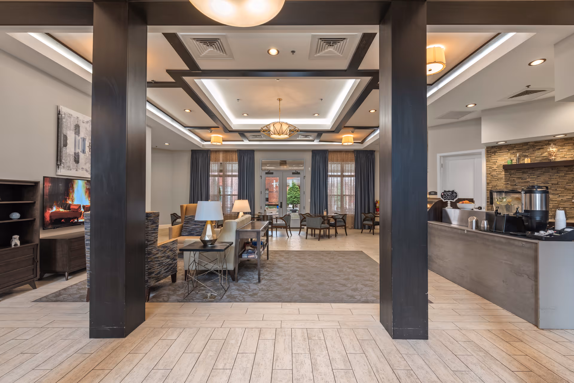 A spacious and well-lit common area in a senior living facility featuring comfortable seating with armchairs and sofas arranged around a coffee table, a flat-screen TV displaying a fireplace scene, large windows with curtains, and a coffee station with various beverage dispensers and cups on the right side.
