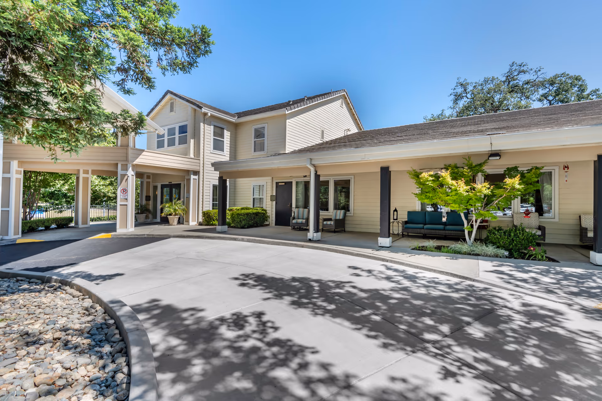 Exterior view of a senior living facility with beige siding and a covered entrance area. There are several chairs and small tables arranged under the covered porch, with some greenery and a small tree planted nearby. The driveway is paved and curves around a landscaped area with rocks and trees casting shadows on the ground. The sky is clear and blue.