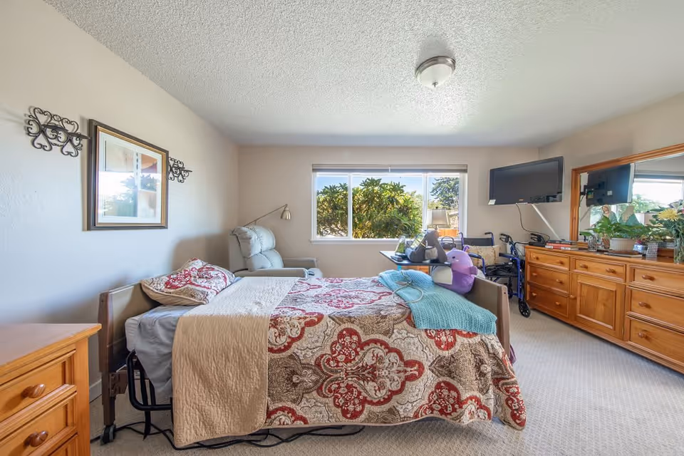 A bedroom in a senior living facility featuring a bed with a patterned quilt, a reclining chair, a wooden dresser with a large mirror, a wall-mounted TV, and a window showing greenery outside. There is a walker next to the dresser and a small table with personal items near the bed.