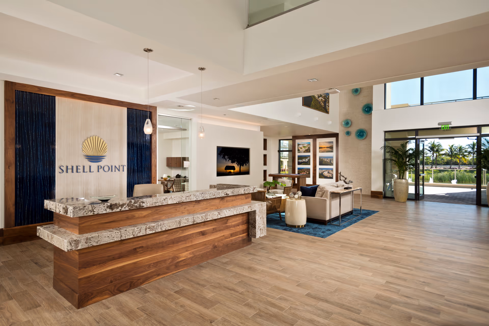 A spacious and modern welcome center interior with a large wooden reception desk topped with granite. Behind the desk is a wall featuring the Shell Point logo. To the right, there is a seating area with sofas, a blue rug, and decorative wall art. Large windows and glass doors allow natural light to fill the space and provide a view of palm trees outside.