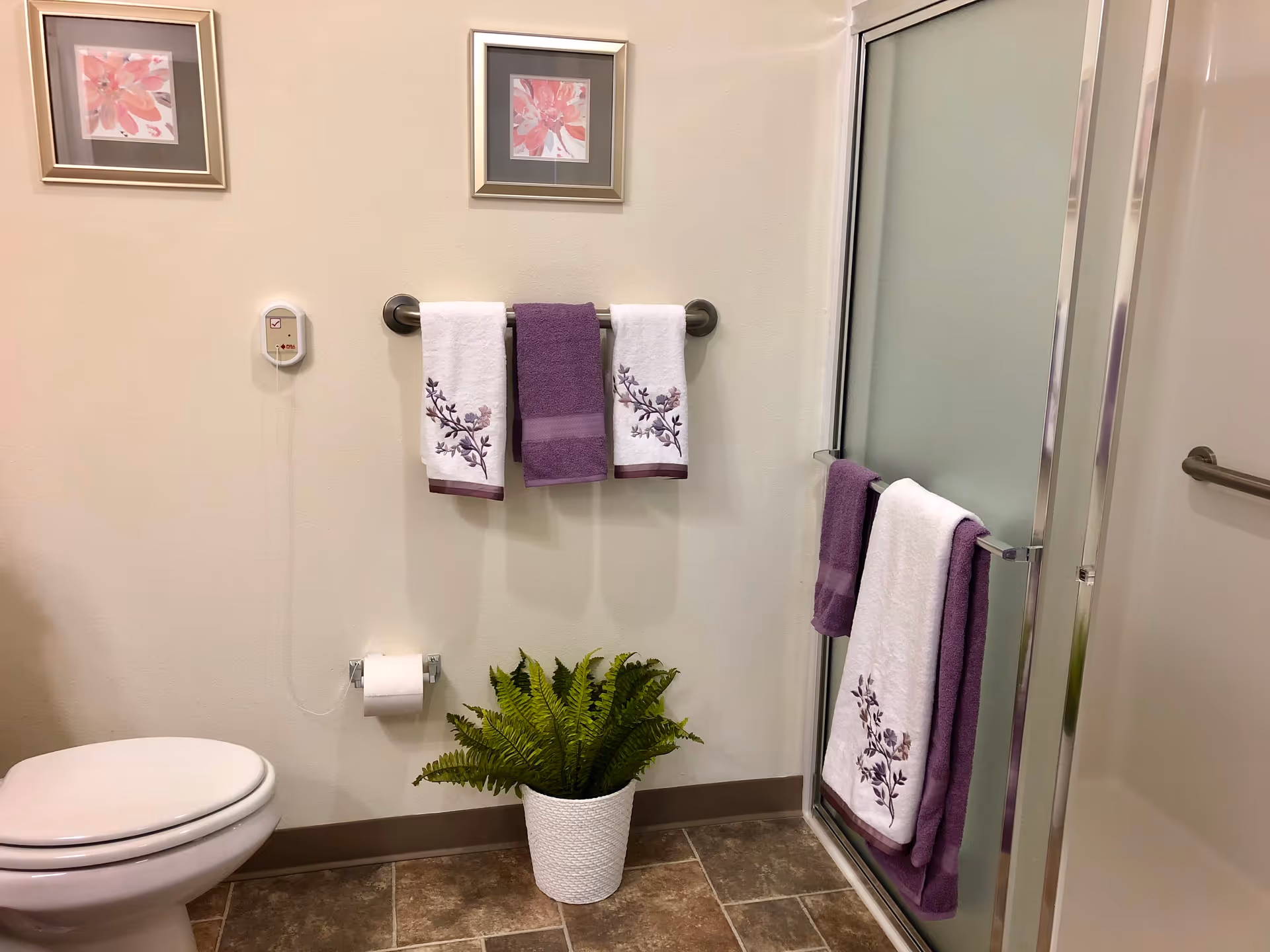 A bathroom with a white toilet on the left, a towel rack on the wall holding two white towels with purple floral designs and one solid purple towel in the middle. Below the towel rack is a toilet paper holder with a roll of toilet paper and a white woven basket with a green fern plant. To the right is a glass shower door with a towel bar holding a white towel with purple floral designs and a solid purple towel. The walls are light-colored, and two framed floral artworks hang above the towel rack.