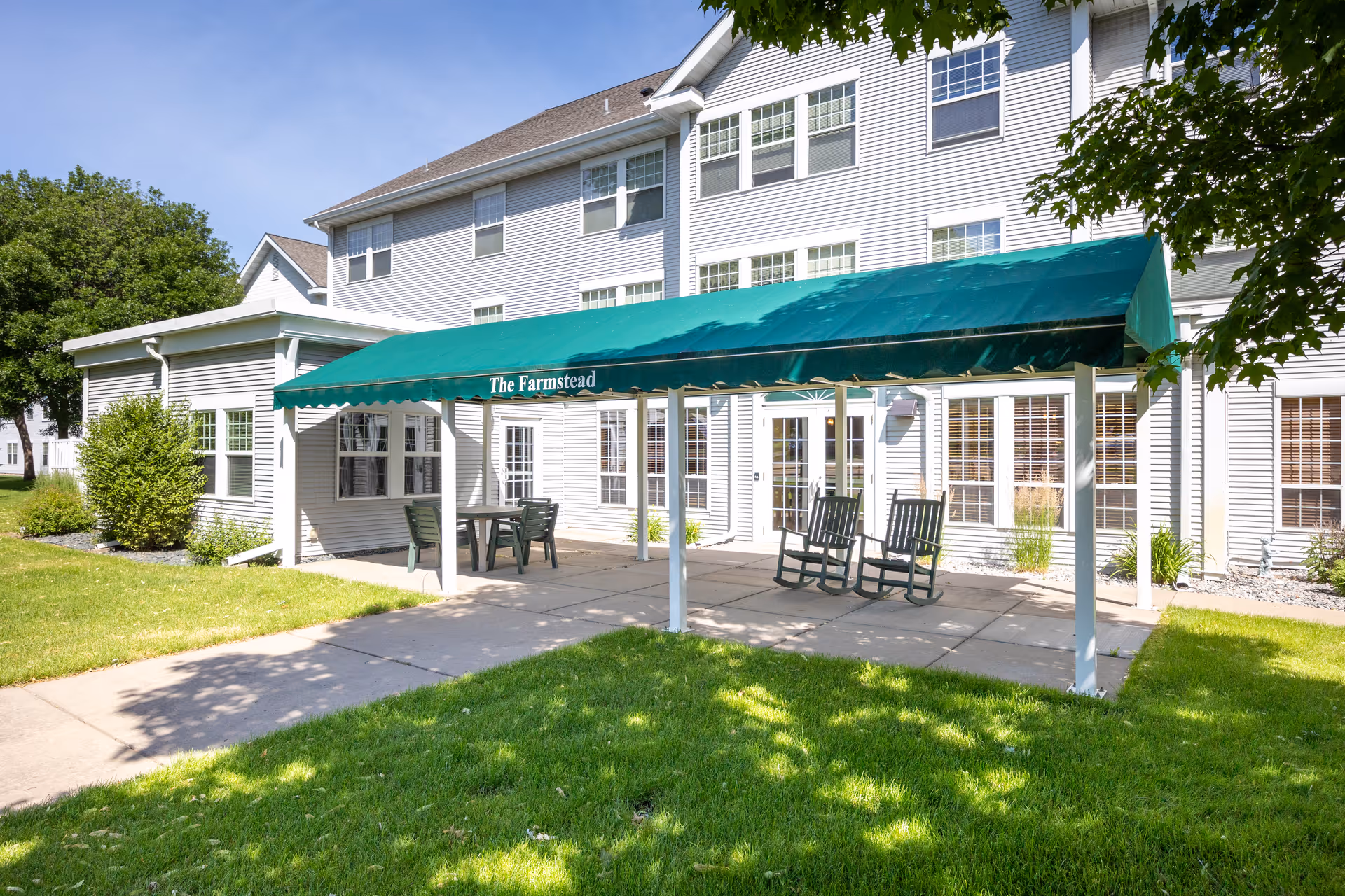 Outdoor patio area at The Farmstead with a green canopy covering a concrete seating area that includes two rocking chairs and a table with chairs. The building behind is a multi-story white structure with many windows and surrounded by green grass and trees.