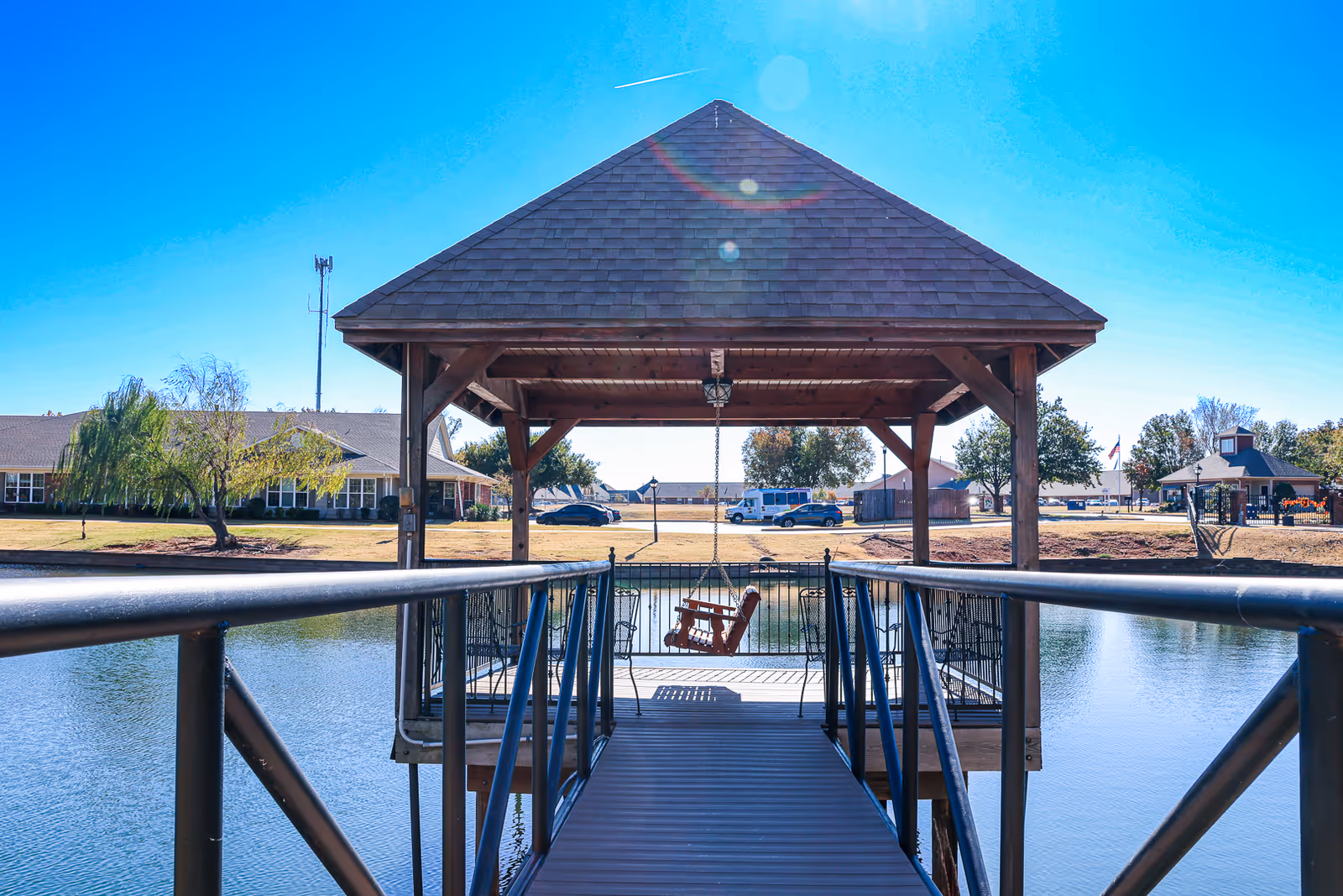 A wooden gazebo with a shingled roof sits at the end of a metal walkway over a pond. Inside the gazebo, a wooden swing hangs from the ceiling. In the background, there are buildings, trees, parked cars, and a clear blue sky.