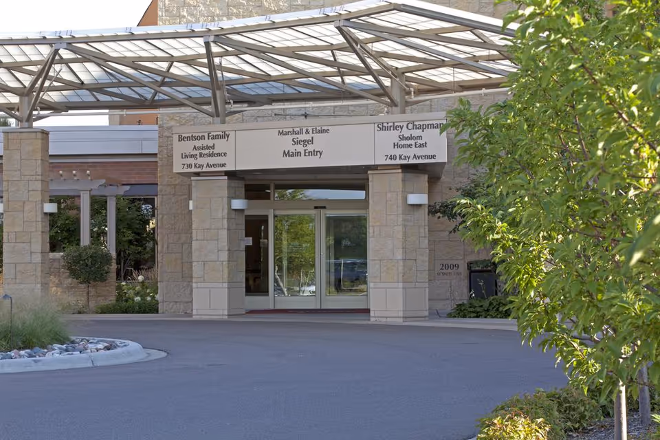 Entrance to a senior living facility with a covered driveway and stone pillars. Signs above the entrance indicate Bentson Family Assisted Living Residence at 730 Kay Avenue, Marshall & Elaine Siegel Main Entry, and Shirley Chapman Sholom Home East at 740 Kay Avenue. There are trees and landscaping around the driveway.