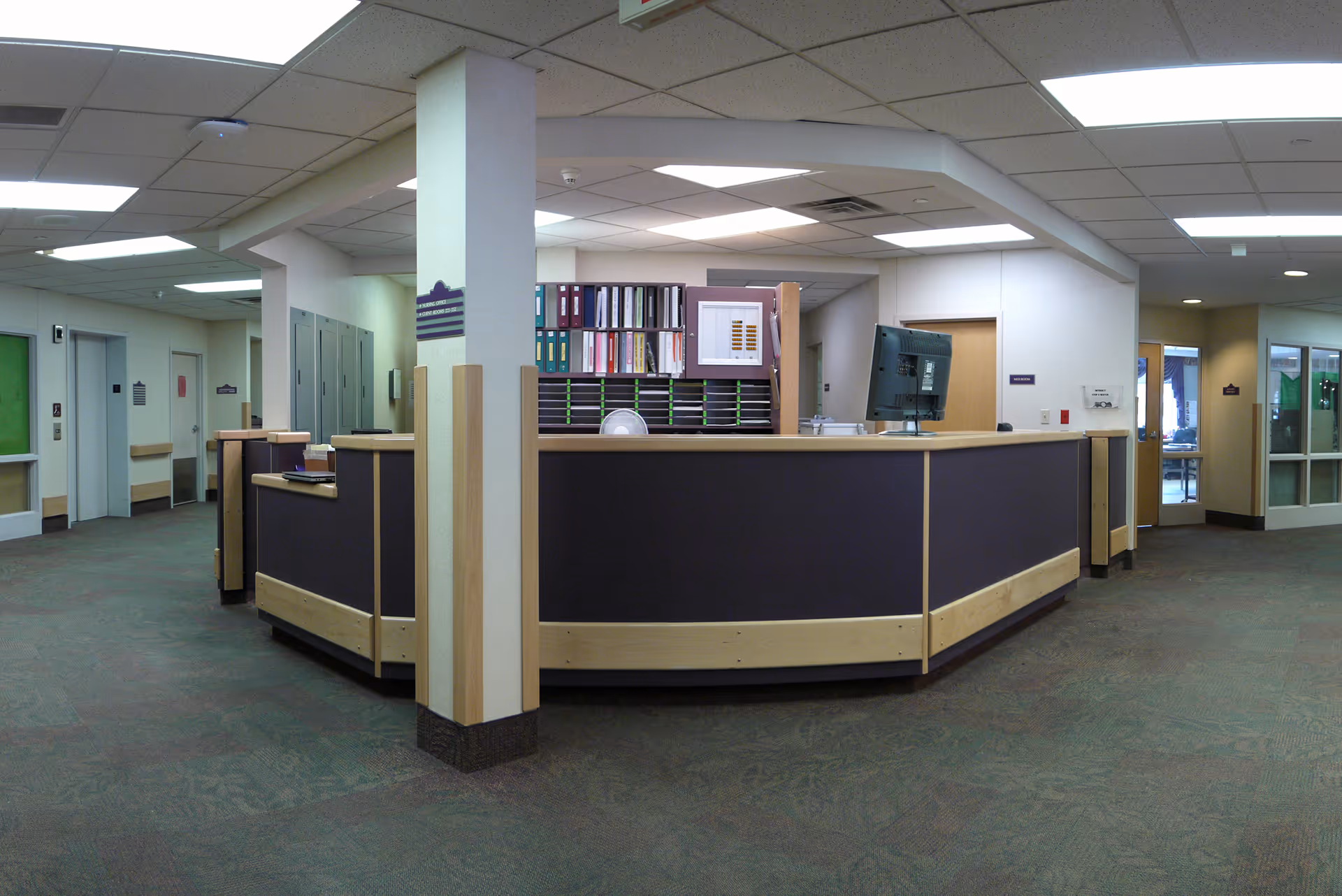 Reception desk area inside a senior living facility with a computer monitor, organized shelves with binders, and multiple doors leading to other rooms. The space is well-lit with ceiling lights and has carpeted floors and beige walls.