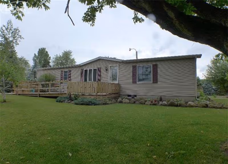 Single-story beige mobile home with maroon shutters, a wooden ramp and deck, surrounded by green grass and trees under a cloudy sky.