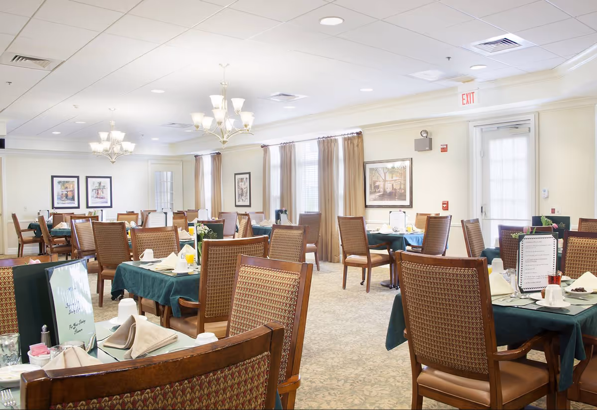 A bright and spacious dining room with multiple tables covered in dark green tablecloths, each set with napkins, menus, and dining ware. The room features wooden chairs with patterned upholstery, large windows with beige curtains, framed artwork on the walls, and elegant chandeliers hanging from the ceiling.