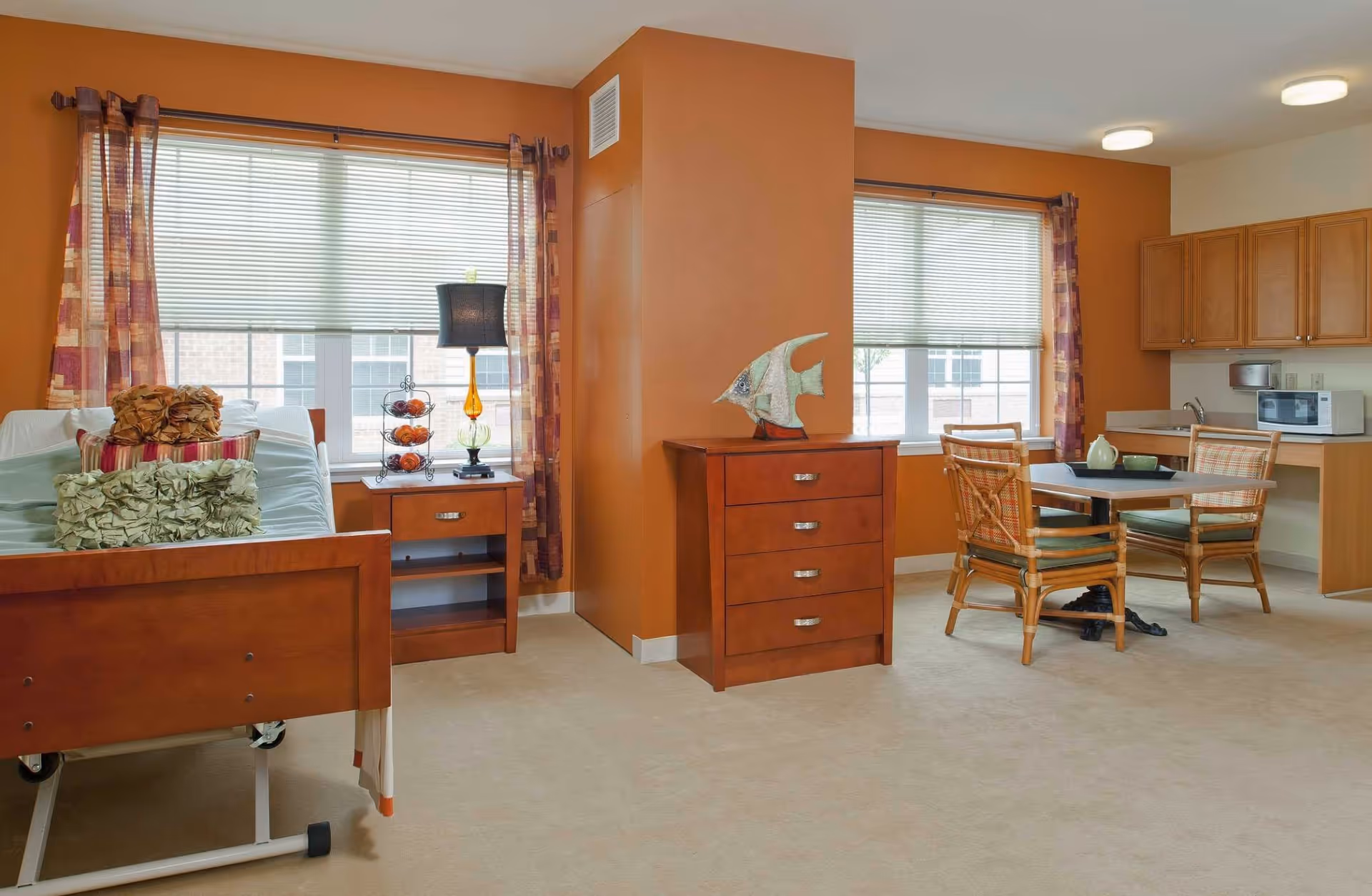 A cozy senior living room with orange walls, two large windows with blinds and patterned curtains, a wooden hospital-style bed with decorative pillows, a wooden nightstand with a lamp and a fruit stand, a wooden chest of drawers with a decorative fish sculpture, a small dining table with four wicker chairs, and a kitchenette area with wooden cabinets, a microwave, and a sink.