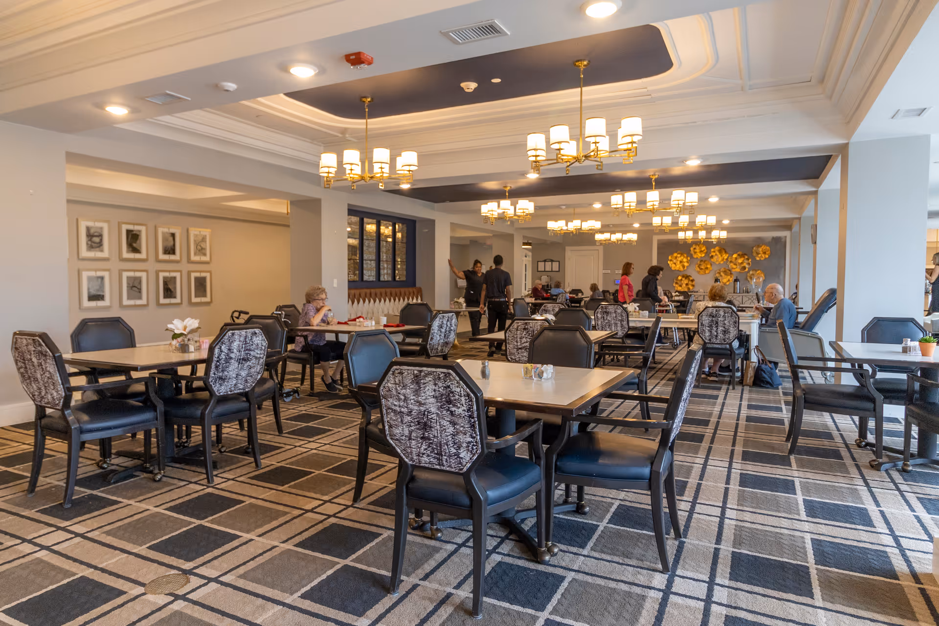 Spacious dining room with multiple tables and chairs, chandeliers overhead, and residents and staff interacting.