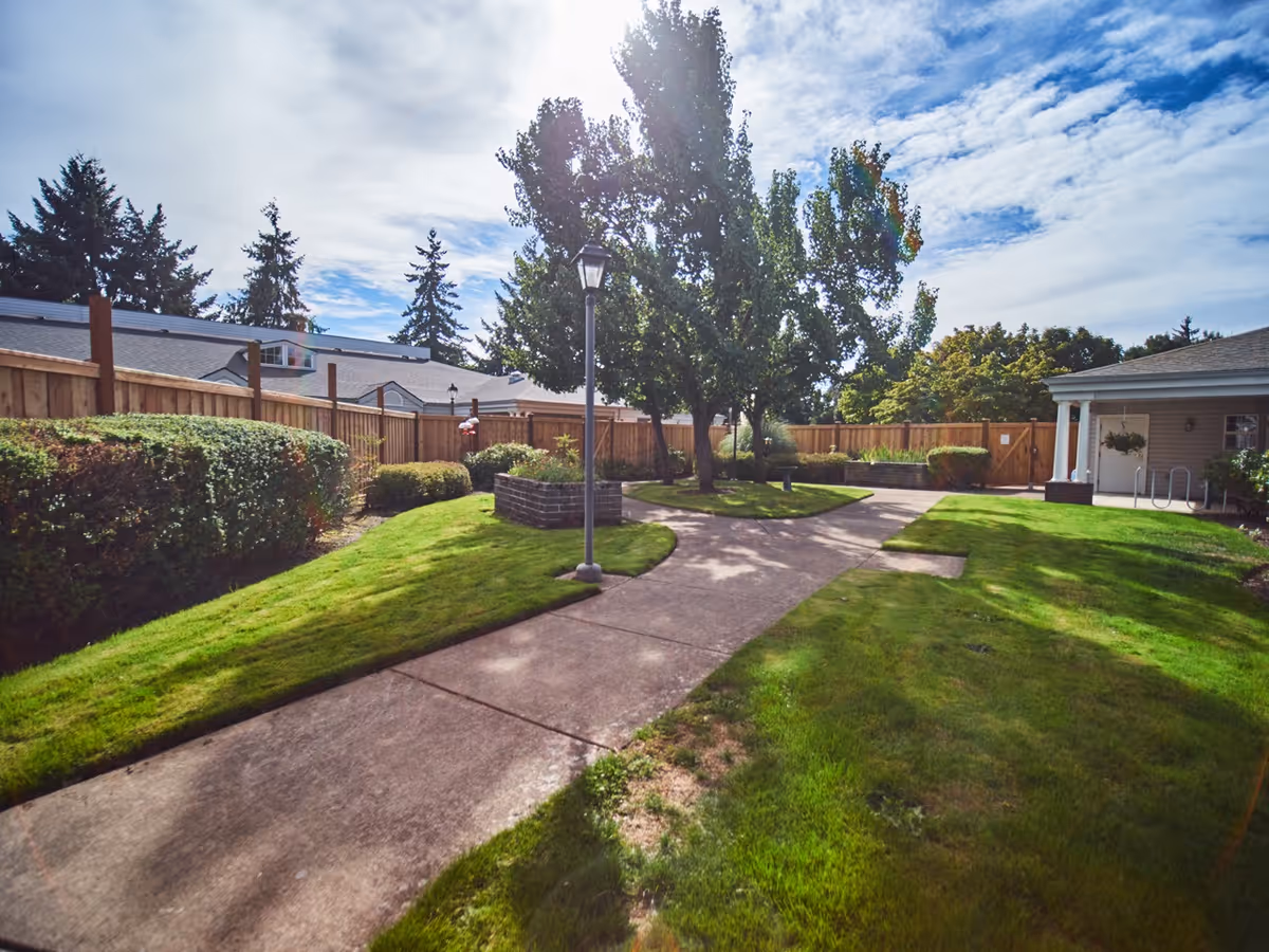 Sunny courtyard with paved walkways, green lawns, trees, a lamppost, and a fenced perimeter beside a single-story building.