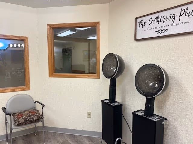 A small room with two black hair drying stations, a gray chair with a patterned cushion, two windows with wooden frames, and a wall sign that reads 'The Gathering Place: sit long • talk much • laugh often.'