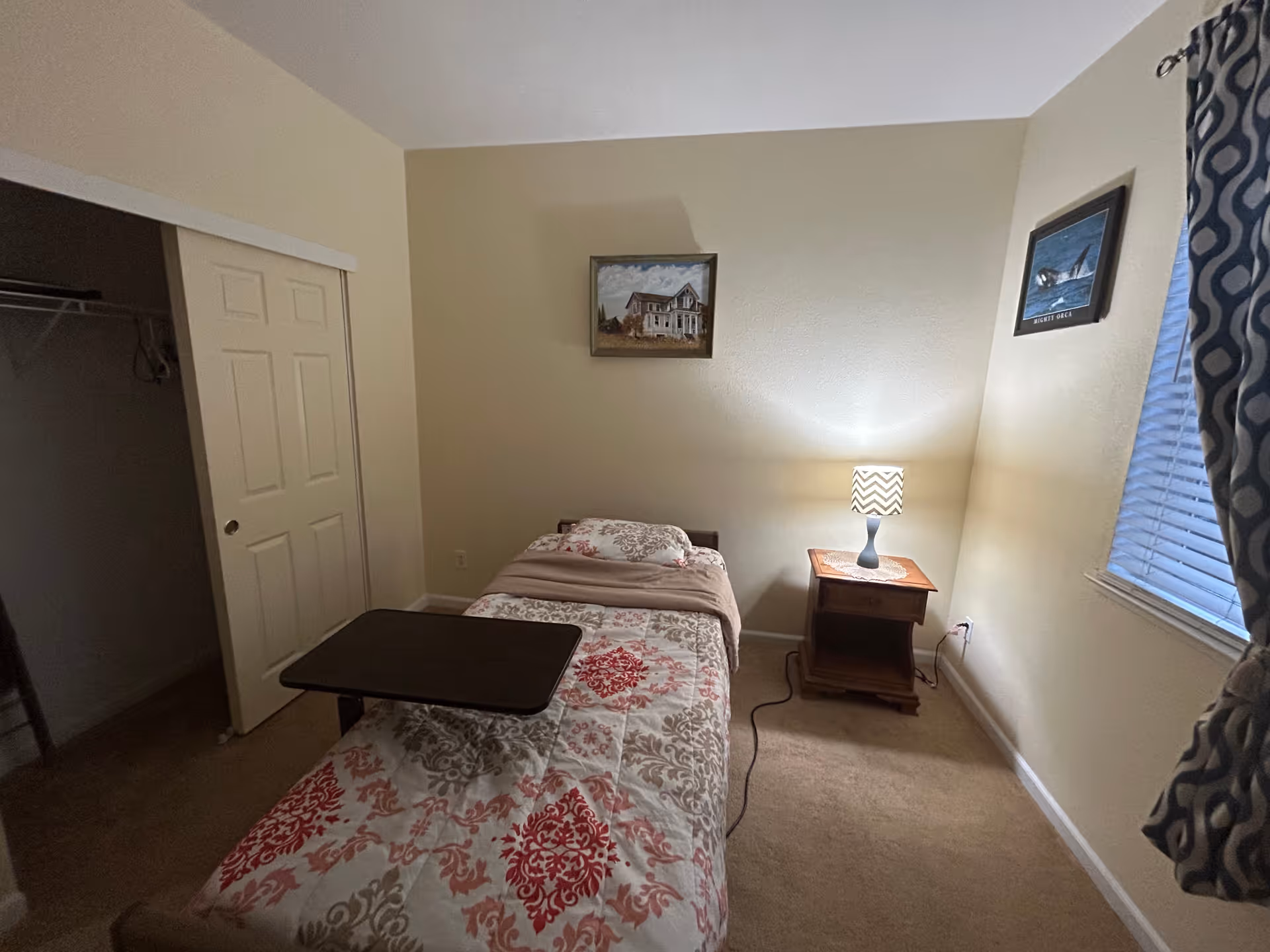 A small bedroom with a single bed covered in a patterned bedspread in red, gray, and white. There is a wooden bedside table with a lamp that has a chevron-patterned shade. The room has beige walls, a window with blue patterned curtains, and two framed pictures on the walls. A closet with sliding doors is visible on the left side.