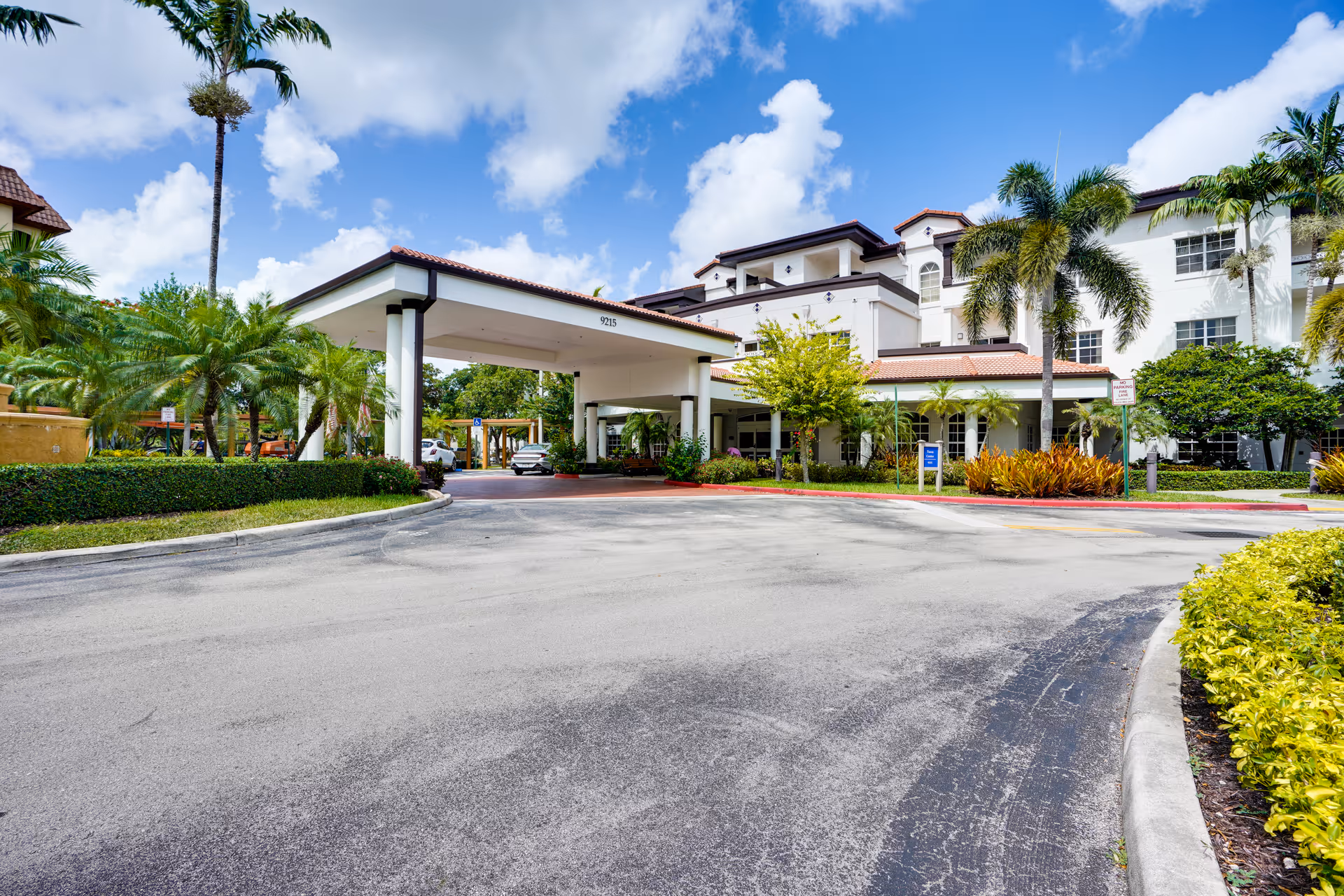Exterior view of a senior living facility with a covered entrance driveway, palm trees, and well-maintained landscaping under a partly cloudy sky.