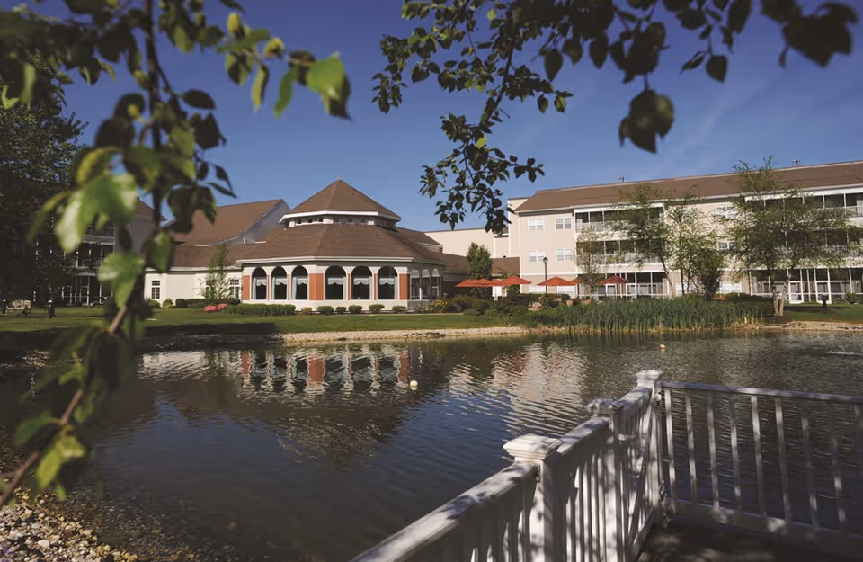 View of a senior living facility named Jefferson's Ferry with a pond in the foreground, a white wooden fence, and buildings with multiple windows and balconies surrounded by greenery under a clear blue sky.