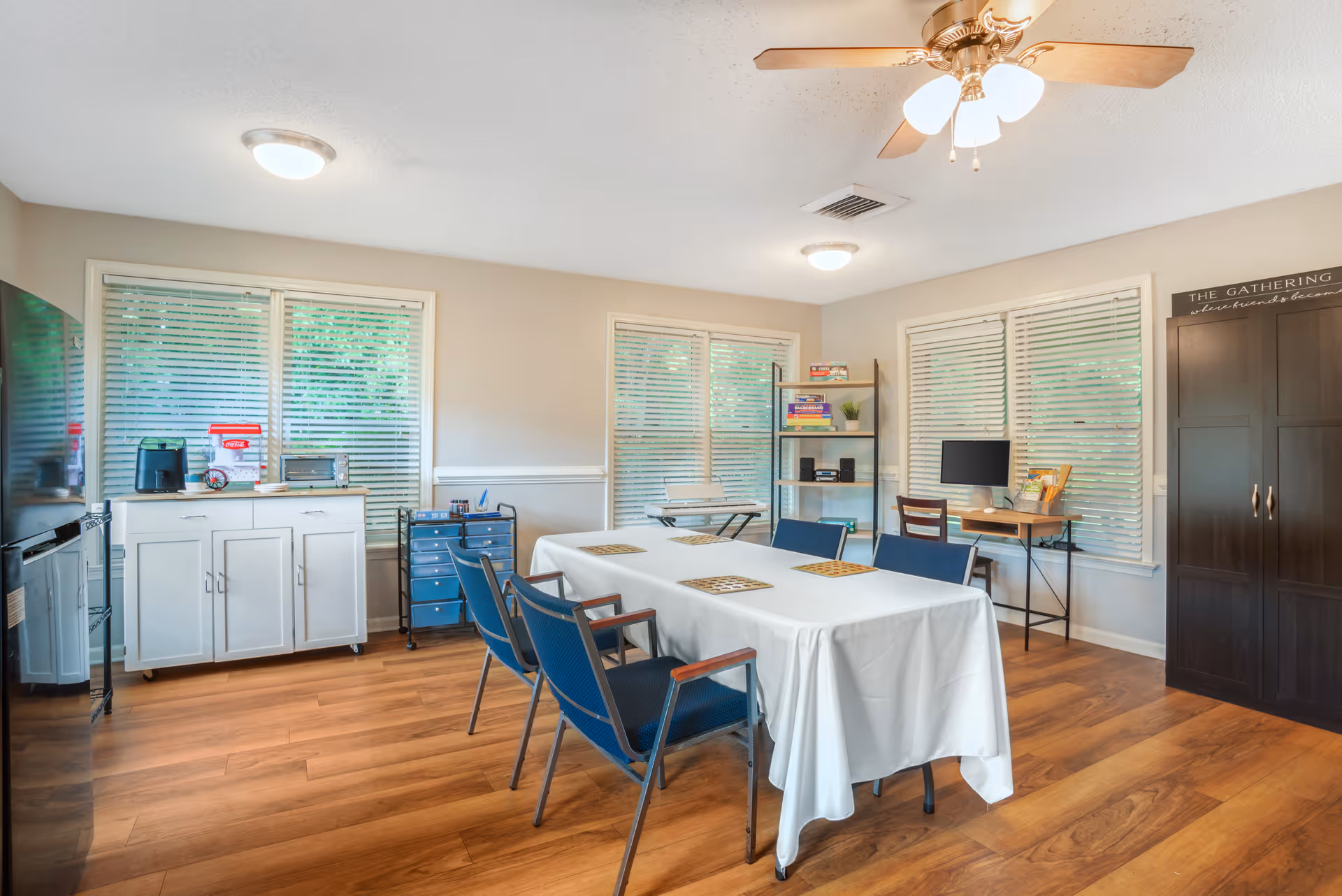 Communal dining/activity room with a long table draped in a white cloth surrounded by blue chairs, cabinets, shelving, and windows with blinds.