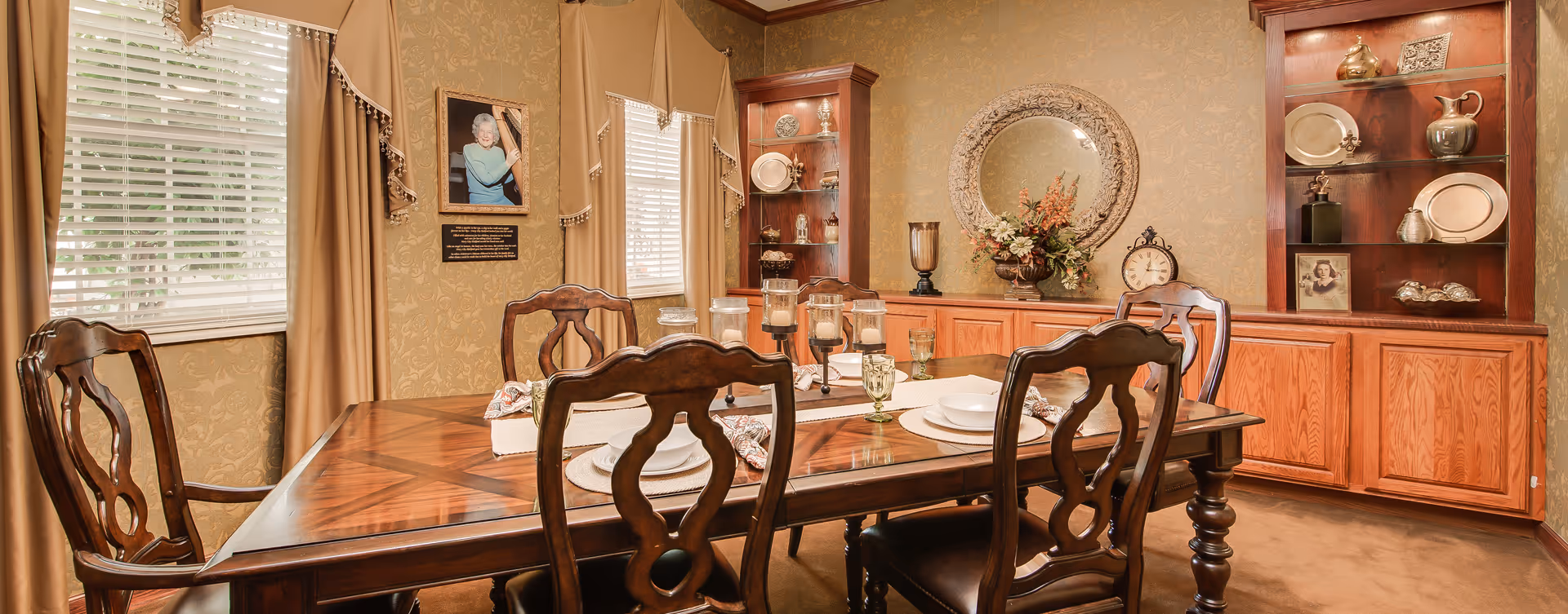 A traditional dining room with a wooden dining table set for four, featuring white plates, bowls, and patterned napkins. The room has beige patterned wallpaper, two windows with beige curtains, and wooden cabinets with glass shelves displaying decorative items. A round ornate mirror, a floral arrangement, and a clock are on the back wall.