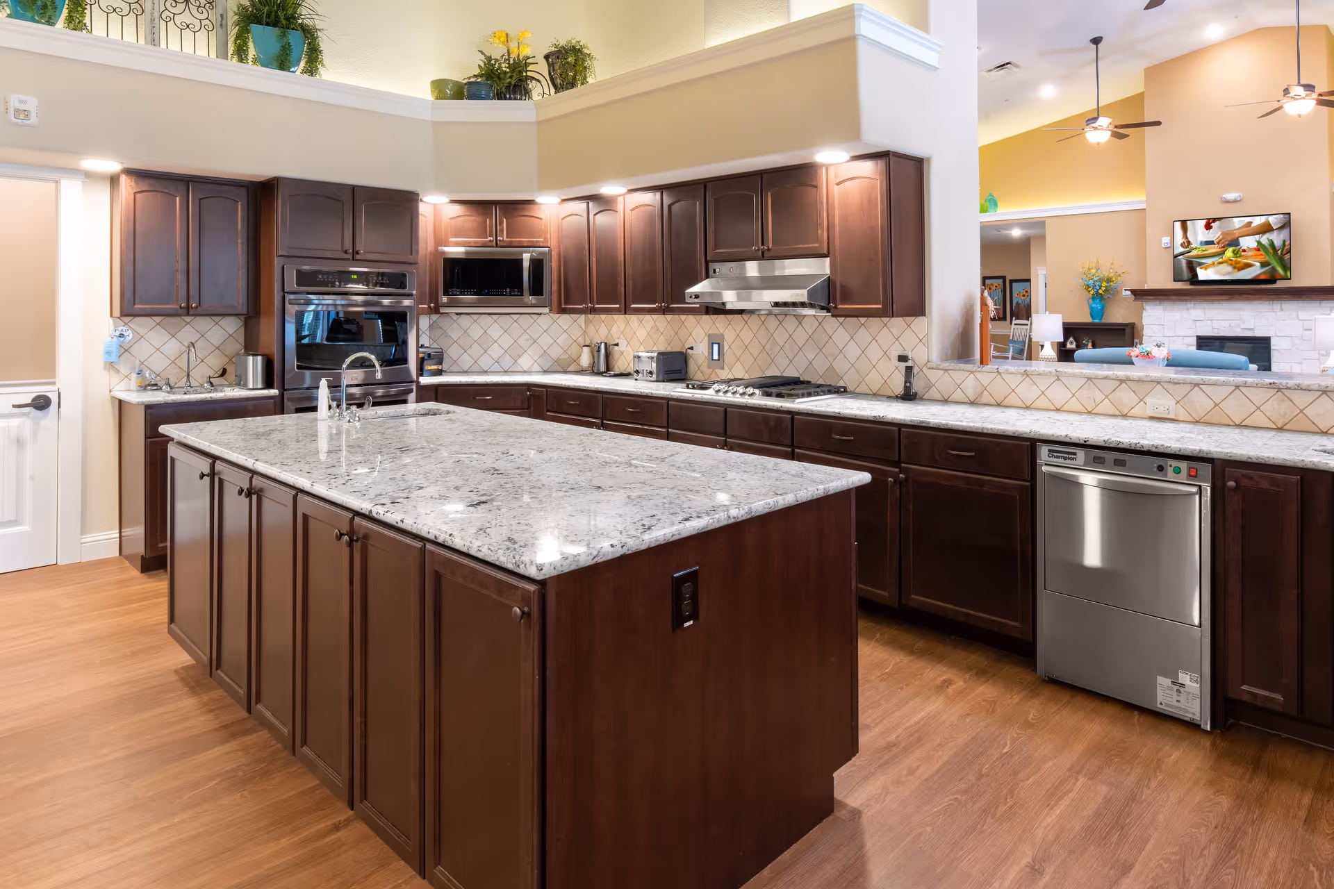 A spacious kitchen with dark wood cabinets and a large central island topped with white and gray granite. The kitchen features stainless steel appliances including a dishwasher, oven, microwave, and stovetop. The backsplash is tiled in a diamond pattern, and the floor is a warm wood tone. In the background, there is a glimpse of a living area with a fireplace, ceiling fans, and decorative plants.