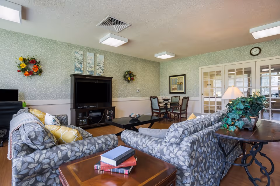 A cozy living room area in a senior living facility with two patterned sofas facing a TV on a wooden stand. There is a coffee table between the sofas and a side table with books and a potted plant. The walls are decorated with floral wreaths and framed artwork. In the background, there is a small dining table with four chairs and French doors leading to another room.