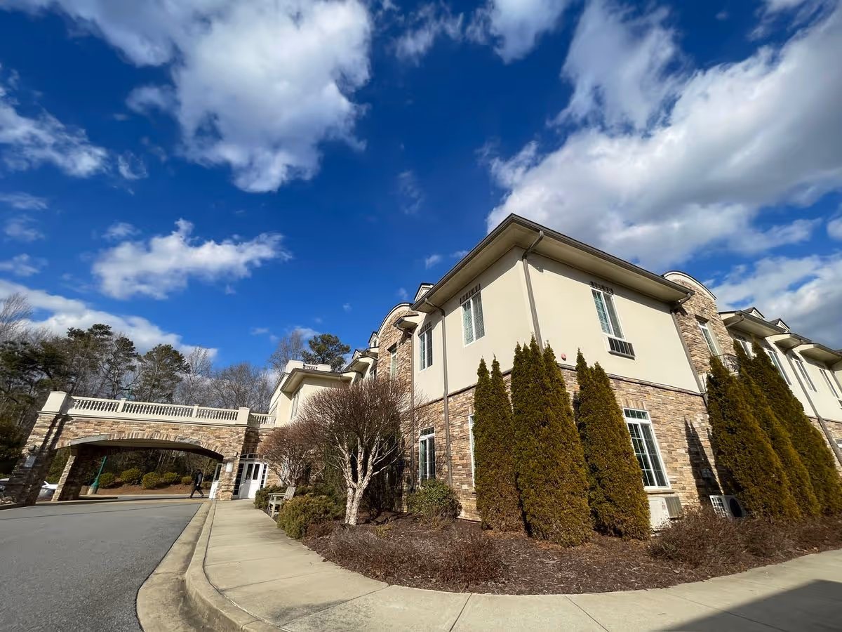 Front exterior of a multi-story senior living building with a stone facade, arched drive-through entrance, and tall shrubs under a blue sky.