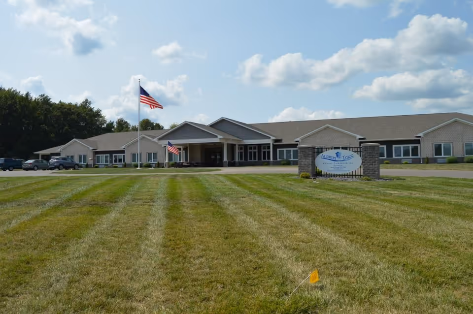 Front view of a single-story senior living building with a wide mowed lawn, flagpoles flying American flags, cars near the entrance, and an 'Autumn Trace' sign.