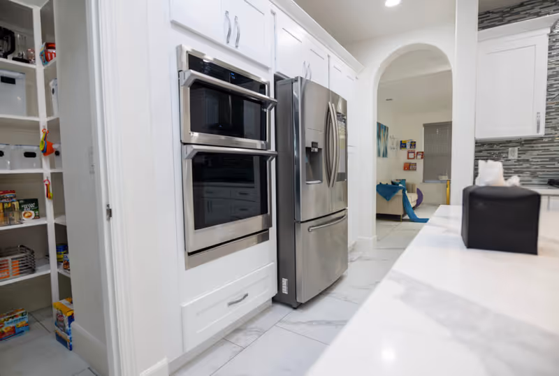 Modern white kitchen with stainless steel double ovens and refrigerator, a marble countertop and an open pantry.